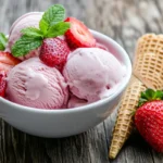 A vibrant bowl of homemade strawberry ice cream, topped with fresh strawberries and mint leaves, set against a rustic wooden background. The ice cream is creamy and smooth, with a rich pink color, and a scoop is elegantly placed on a waffle cone beside it. Soft natural light enhances the textures and colors, creating a mouth-watering appeal.